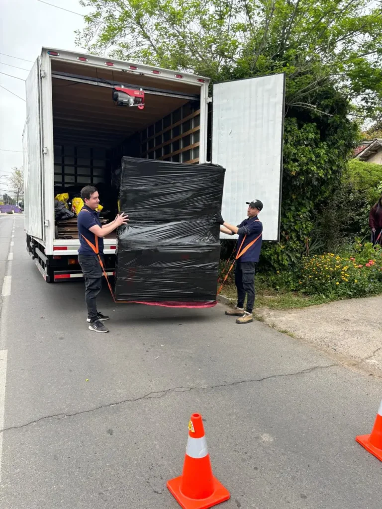 Dos trabajadores de mudanza cargando muebles envueltos en plástico negro desde camión blanco en calle residencial